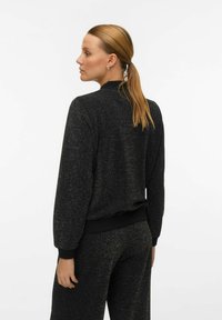 Veste noire texturée au design sportif, avec des poignets et un ourlet côtelés. Des paillettes dorées créent un effet scintillant subtil.