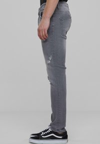 Szare wąskie dżinsy z przetarciami na udzie, pięciokieszeniowy krój i lekko wyblakłe wykończenie, w zestawieniu z czarnymi trampkami.