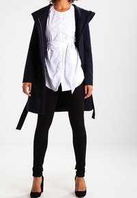 Navy long coat with hood, paired with a white button-up shirt and black leggings. Features a zip closure and belted waist. Black heels complete the look.