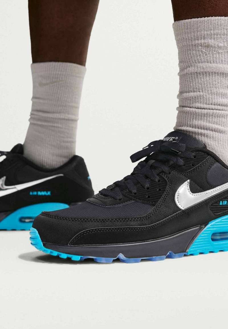 Sorte og blå Nike Air Max sneakers båret med hvide sportssokker, vist på fødder mod en neutral baggrund.