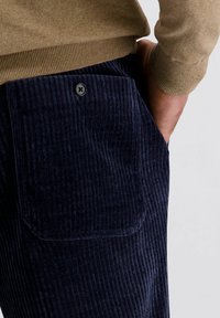 Man in een marineblauwe corduroy broek met een knoopzak aan de achterkant en een camelkleurige trui, hand in zijn zijzak.