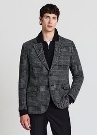 Blazer a quadri in bianco e nero, doppiopetto con rever a lancia e chiusura con bottoni, indossato sopra una polo a maglia scura. Tessuto in lana strutturata.