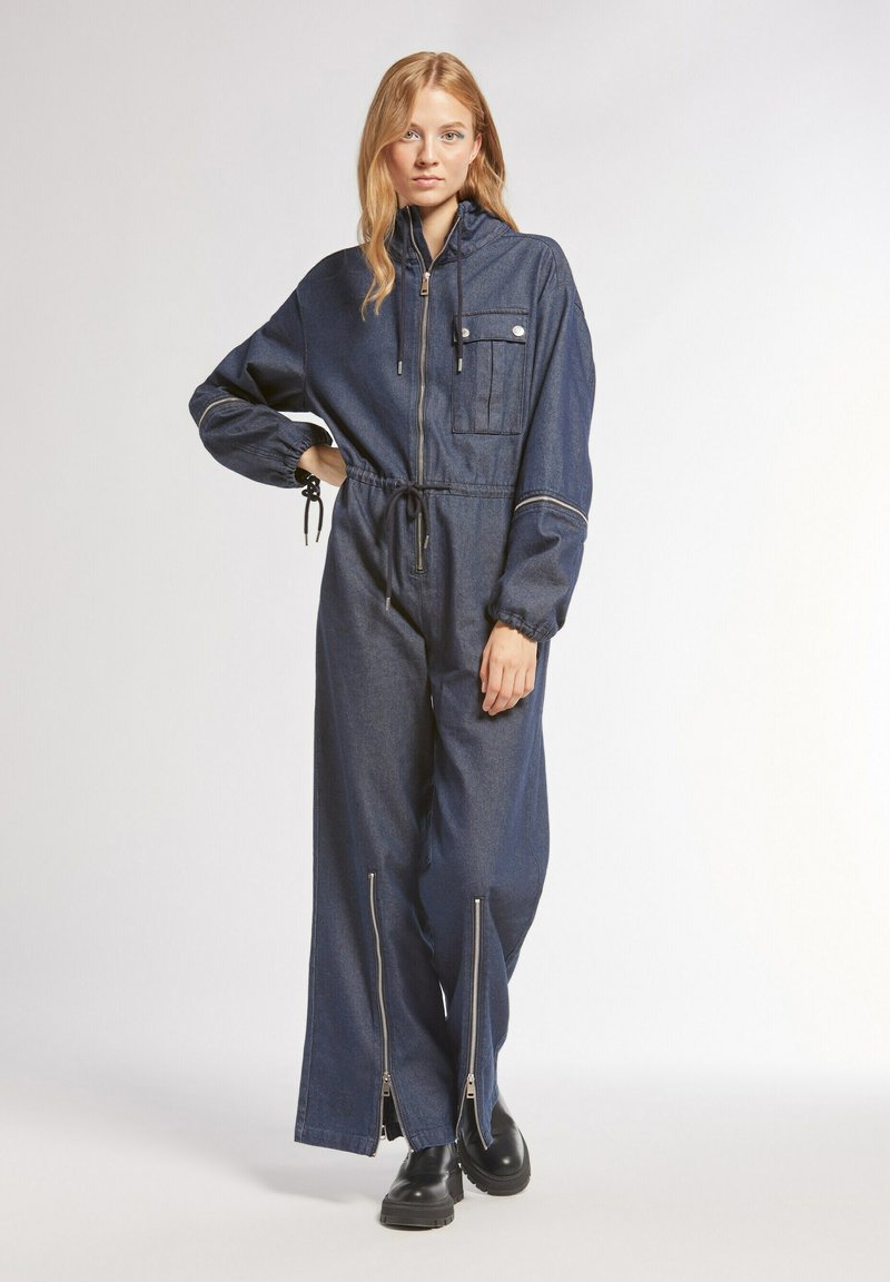 Salopette in denim con zip frontale, cappuccio e tasca sul petto. Presenta maniche lunghe, polsini elasticizzati e zip sui polpacci. Colore: blu scuro.