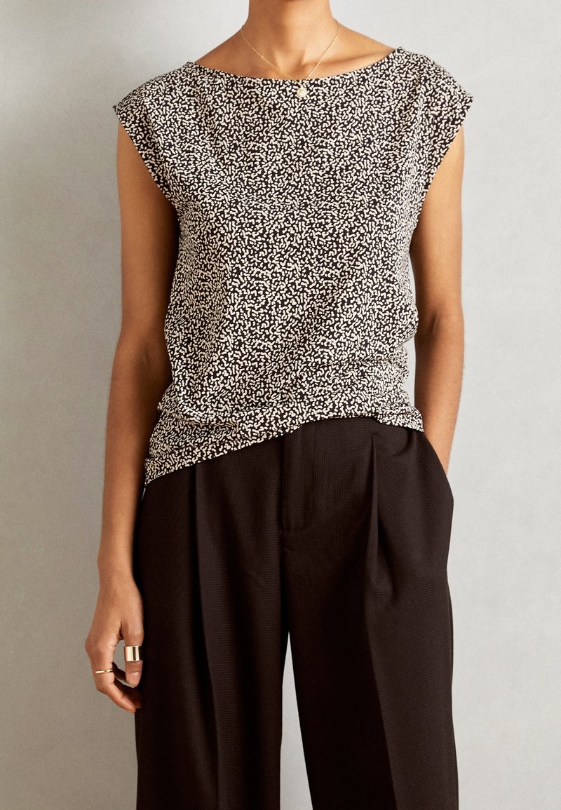 Femme portant un haut sans manches à motifs noirs et blancs, rentré dans un pantalon noir ample, main dans la poche et bagues en or.