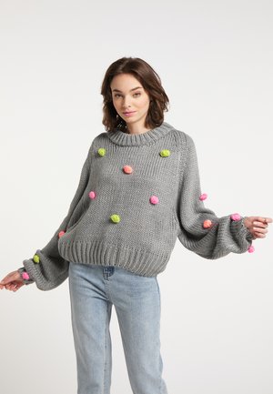 Jonge vrouw die een losse grijze gebreide trui met kleurrijke pompons en lichtblauwe spijkerbroek draagt, staand tegen een egale lichte achtergrond.