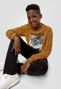 Musztardowy sweter z nadrukiem graficznym, czarne spodnie i białe sneakersy. Materiał wygląda na miękki, z dopasowanymi mankietami i luźnym krojem.