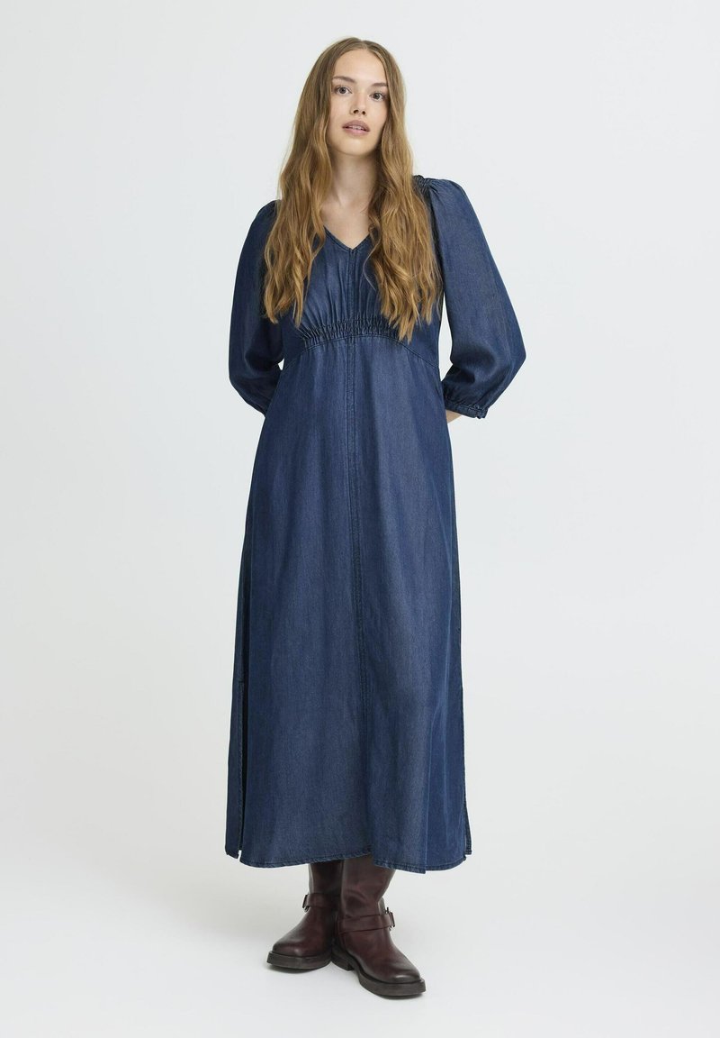 Langdenim jurk met een V-hals en geplooide details op de taille, voorzien van lange, opgeblazen mouwen en een vloeiende silhouet, gecombineerd met bruine laarzen.