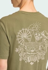 Olivovo zelené bavlnené tričko s detailnou bielou grafickou potlačou rôznych ikon, vrátane ruského kola, akordeónu a preclíka.