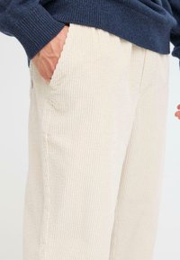 Beige fløjlbukser med en ribbet struktur og sidelommer, bæres med en mørkeblå, strikket sweater, der er delvist synlig.