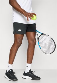 Tennisshorts i sort med hvid Adidas-logo, parret med en hvid t-shirt, sorte træningssko, hvide sokker og holder en grøn tennisbold.