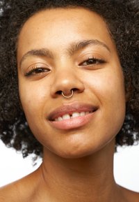 Nahaufnahme einer Person mit kurzem, lockigem schwarzen Haar und einem goldenen Septumring. Die Haut ist glatt mit einem natürlichen Glow. Einfacher Hintergrund.