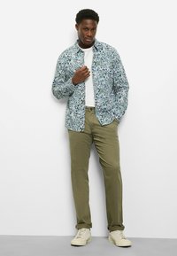 Langärmliges Hemd mit blau-grünem Blumenmuster, kombiniert mit olivgrünen Chinos und cremefarbenen Sneakers. Das Modell steht lässig mit einer erhobenen Hand.