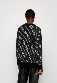 Zwarte gebreide trui met een pluizige textuur, oversized pasvorm en herhaald "HUGO" logo in wit. Geribbelde manchetten en zoom, met een ronde hals.