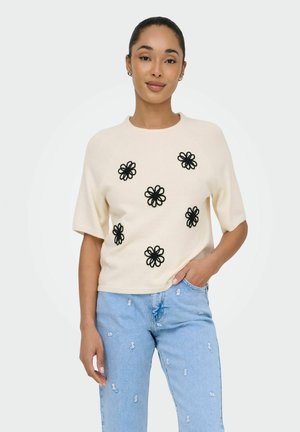 Vrouw die een crèmekleurige trui met korte mouwen draagt met zwarte bloemapplicaties en lichtblauwe versleten spijkerbroek, staand met één hand in de zak.