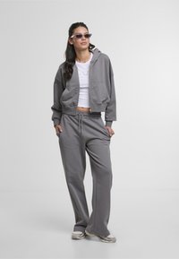 Sweat à capuche gris, court, avec fermeture éclair et poches latérales, associé à un pantalon de jogging gris. Les deux sont fabriqués dans un tissu doux et présentent un design sportif.