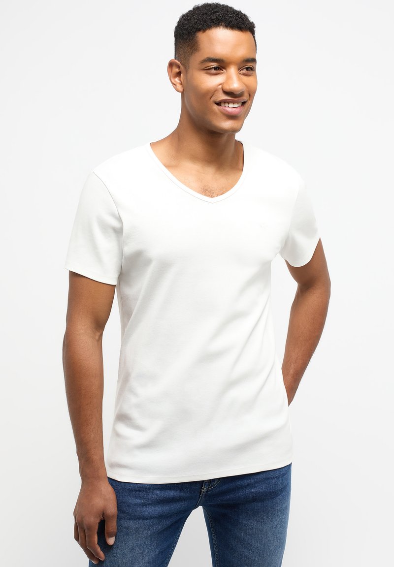 Tricou alb cu decolteu în V, confecționat din bumbac, cu mâneci scurte și o textură netedă, prezentat alături de blugi ajustați de culoare albastră.