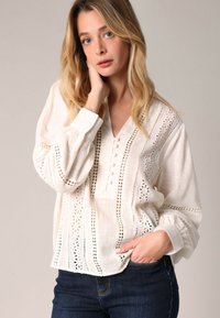 Blouse en lin blanc avec des détails en dentelle ajourée, patte de boutonnage et manches longues. Portée avec un jean en denim foncé, elle présente une coupe décontractée.