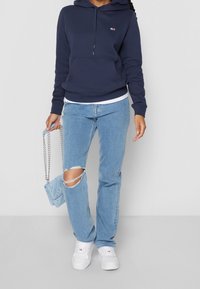 Sudadera con capucha azul marino, vaqueros desgastados de color azul claro, zapatillas blancas y un pequeño bolso azul claro con cadena. Diseño sencillo y ajuste casual.