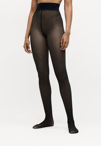 Woman wearing sheer black tights standing barefoot against a plain white background, showing legs and partial torso.