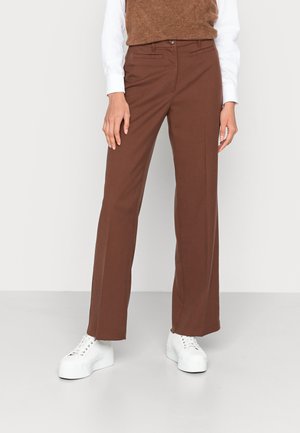 Frau trägt braune gerade geschnittene Hose, weißes Langarmshirt, braunen ärmellosen Pullover und weiße Plateausneaker, steht vor weißem Hintergrund.