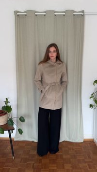 Bēšs mētelis ar kapuci, priekšējo rāvējslēdzēju un jostu pie vidukļa, ar divām sānu kabatām un gludu tekstūru, kombinēts ar melnām plaša piegriezuma biksēm.