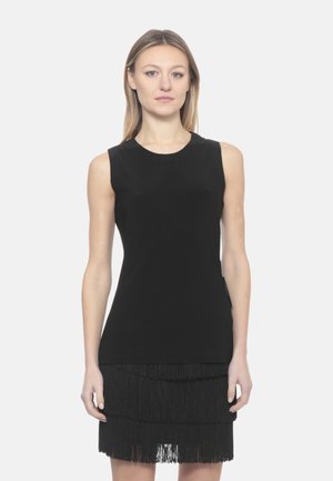 Donna con capelli biondi lisci che indossa un vestito nero senza maniche con gonna a frange a strati, in piedi davanti a uno sfondo bianco uniforme.