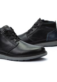 Botas de tobillo de cuero negro con suelas de goma gris, diseño de cordones, acentos azules en el talón, textura suave y costuras mínimas.
