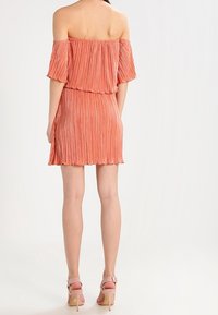 Robe décolletée, texture plissée, en corail clair. Longueur courte, design superposé et ourlet festonné. Portée avec des talons hauts roses assortis.