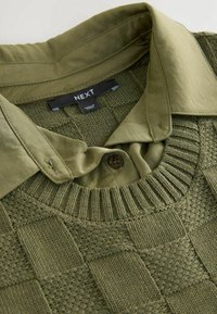 Pull en maille verte avec un col côtelé superposé à une chemise verte boutonnée avec un col pointu et une étiquette de marque "NEXT" à l'intérieur de l'encolure.