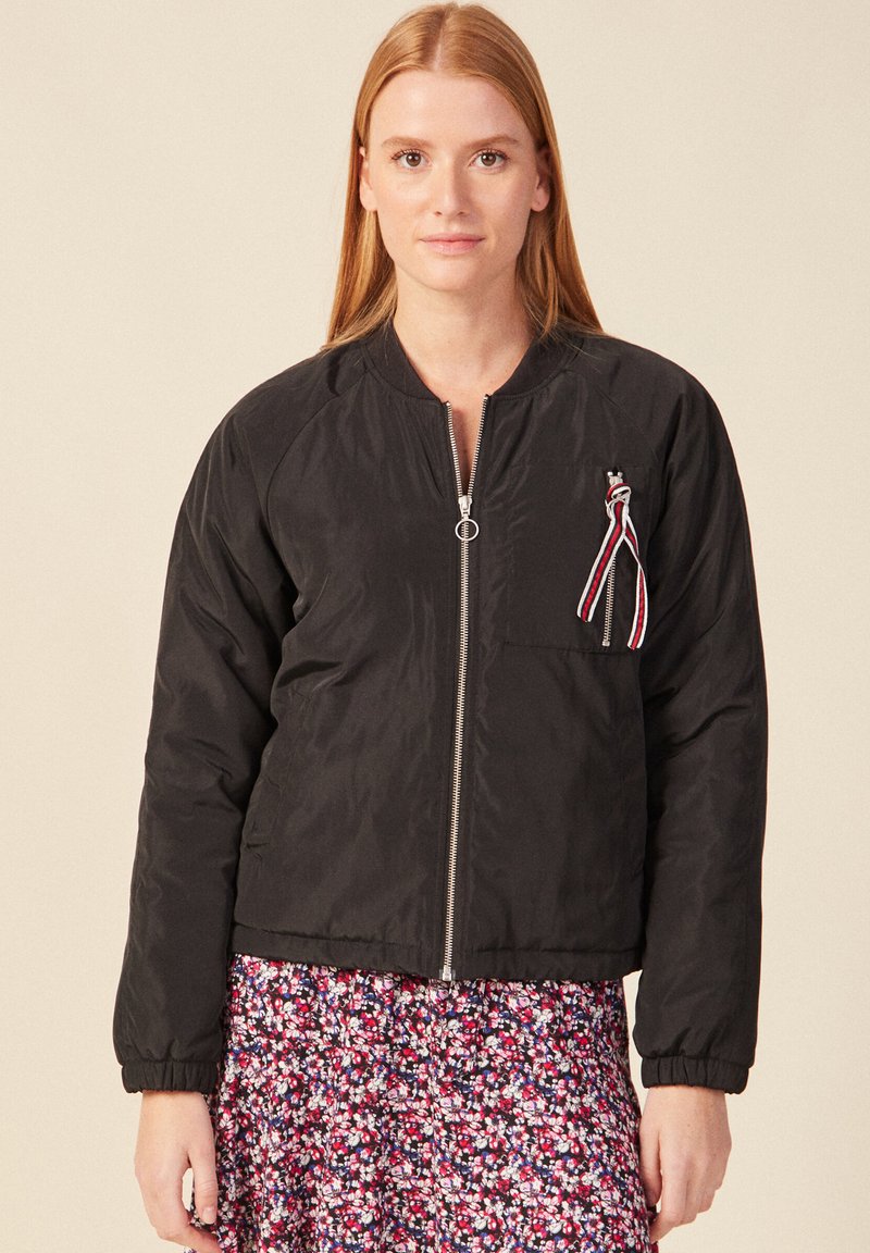 Zwarte bomberjack met een ritssluiting aan de voorkant, geribbelde manchetten en een borstzak met een veelkleurige lintaccent. Soepele textuur.