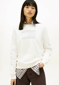 Young woman with straight black hair wears a white Tommy Jeans sweatshirt over a plaid shirt, posing with one hand in dark pants pocket.