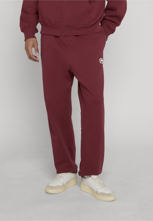 Hombre vestido con pantalones deportivos granate y chaqueta a juego con cremallera, con zapatillas blancas, de pie contra un fondo liso gris claro.