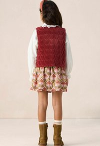 Child wearing a white long-sleeve blouse, red quilted vest with heart patterns, floral skirt, brown boots, and a red headband, facing away.