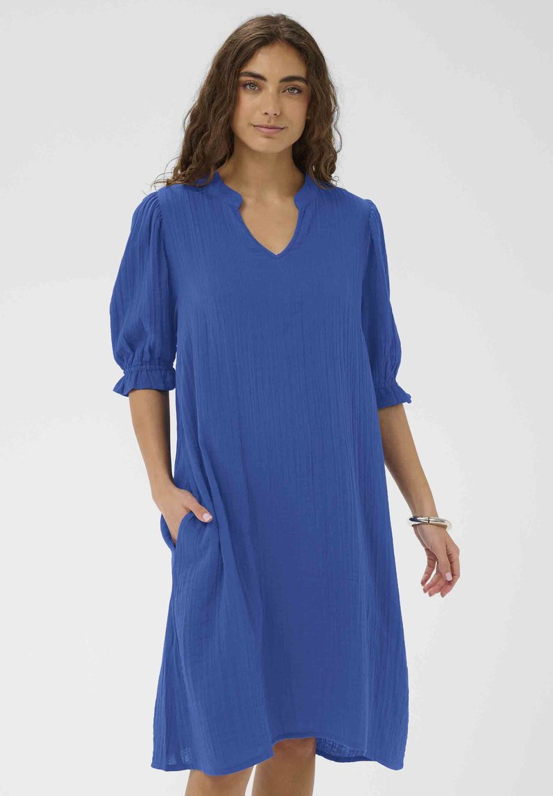 Femme portant une robe bleue ample longueur genou avec manches bouffantes 3/4, encolure en V et poches latérales, debout contre un fond clair uni.