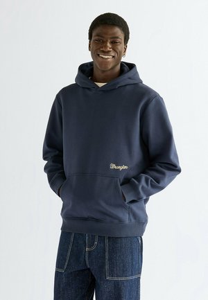 Uśmiechnięty mężczyzna w granatowej bluzie z kapturem Wrangler, z rękami w przedniej kieszeni, w ciemnoniebieskich dżinsach na jednolitym jasnym tle.