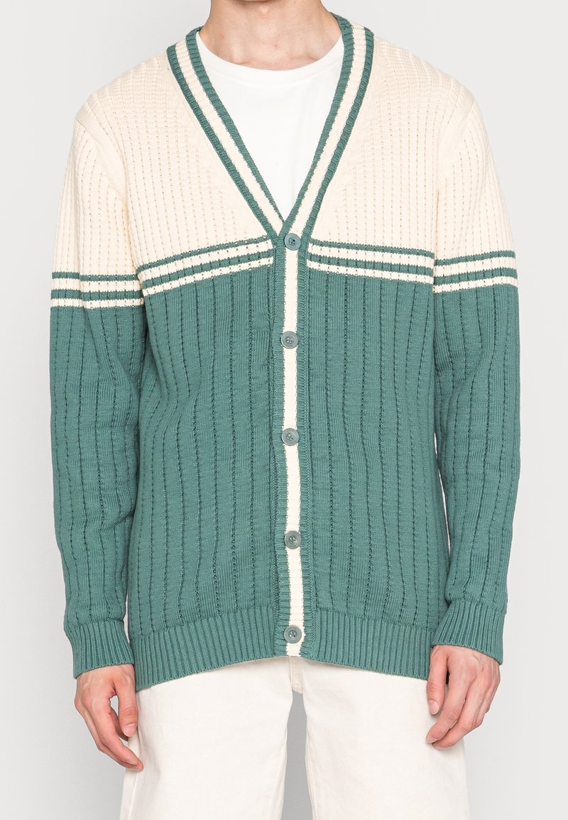 Homme portant un cardigan en tricot boutonné vert et crème avec un motif côtelé vertical, sur un t-shirt blanc et un pantalon clair.