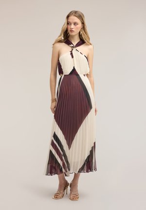 Vrouw in een mouwloze geplooide maxi-jurk met bordeauxrode, zwarte en witte geometrische patronen, draagt gouden sandalen met hoge hak en oorbellen.