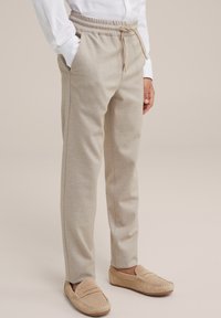 Pantalones jogger de color beige claro, hechos de tela suave, con cinturilla elástica, detalle de cordón y bolsillos laterales. Combinados con zapatos de deslizamiento beige.