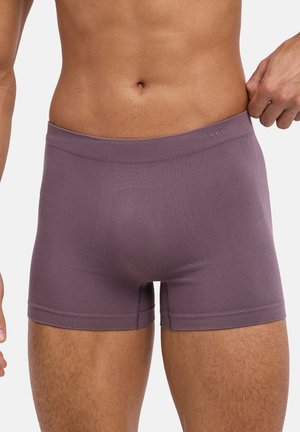 Mannelijk torso met paarse boxershorts, licht de tailleband naar buiten trekkend, waardoor pasvorm en stoftextuur zichtbaar zijn.