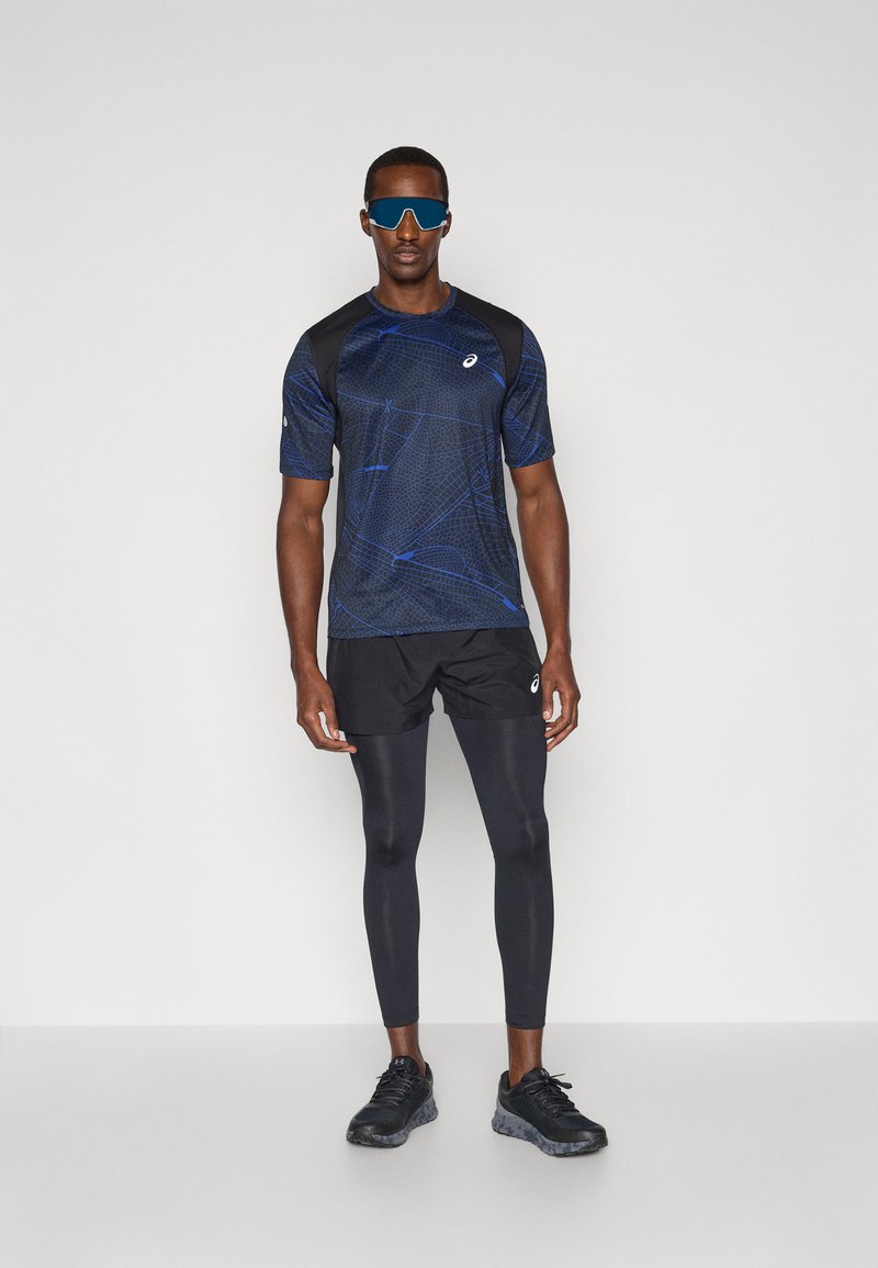 Sportsoutfit bestående af en kortærmet blå mønstret skjorte, sorte shorts, sorte leggins og sorte sneakers. Letvægts- og åndbare materialer.