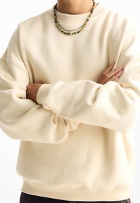 Jasno beżowy sweter z prążkowanym dekoltem i mankietami. Materiał wygląda na miękki i ma luźny krój. Ma małe logo na rękawie.