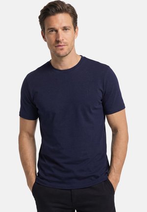 Uomo con capelli corti castani, che indossa una maglietta blu navy a maniche corte e pantaloni neri, in piedi con le mani nelle tasche.