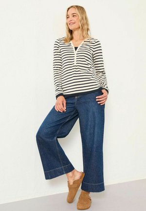 Langärmliges gestreiftes Oberteil in Creme und Marineblau, mit V-Ausschnitt und Knopfdetails, kombiniert mit weit geschnittenen Jeanshosen in dunklem Denim. Weiche, legere Schuhe.