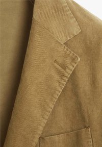 Blazer de pana en marrón claro. Presenta solapas anchas, un bolsillo y una textura de cordones verticales. Acento cosido visible en el cuello.