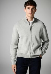 Grau gefütterte Strickjacke mit Reißverschluss, strukturiertem Strick, hohem Kragen und gerippten Bündchen. Getragen über einem weißen Hemd, kombiniert mit dunklen Jeans.