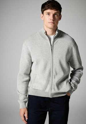Grau gefütterte Strickjacke mit Reißverschluss, strukturiertem Strick, hohem Kragen und gerippten Bündchen. Getragen über einem weißen Hemd, kombiniert mit dunklen Jeans.