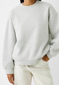 Light grey sweatshirt made of soft fabric, featuring a crew neck, dropped shoulders, and ribbed cuffs and hem. Paired with light pants.