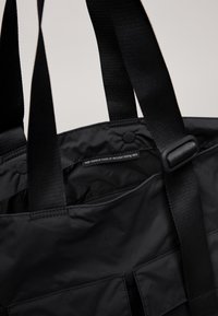 Svart toteväska tillverkad av återvunna fiskenät, med två robusta handtag, en slät yta och en etikett som anger materialets sammansättning.