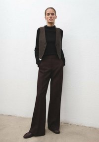 Gilet in lana marrone con bottoni, top a maniche lunghe nero a costine e pantaloni larghi marroni, abbinati a scarpe scure, su uno sfondo di parete bianca semplice.