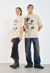 Camisetas oversized beige con estampado abstracto negro, combinadas con jeans acampanados negros y botas de tobillo negras en un modelo, y jeans azules en el otro.